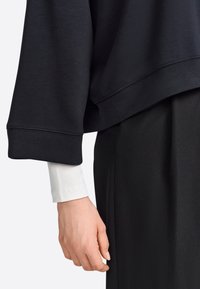 Svart huvtröja med breda, korta ärmar över en vit långärmad topp. Slät tygkvalitet med rena sömsdetaljer.