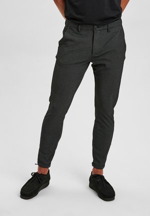 Mężczyzna ubrany w dopasowane, ciemnoszare spodnie w wzory, czarne buty i czarną koszulę, stojący na tle jednolitego, jasnego tła.