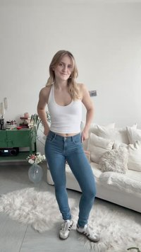 Ung kvinde iført hvidt tanktop og jeans står smilende i lyst opholdsstue med hvid sofa og grønt skænk.