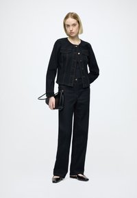 Tmavá rifľová bunda so zlatými gombíkmi, kombinovaná s tmavými džínsami s širokými nohavicami. Modelka drží čiernu tašku s oblým dizajnom.