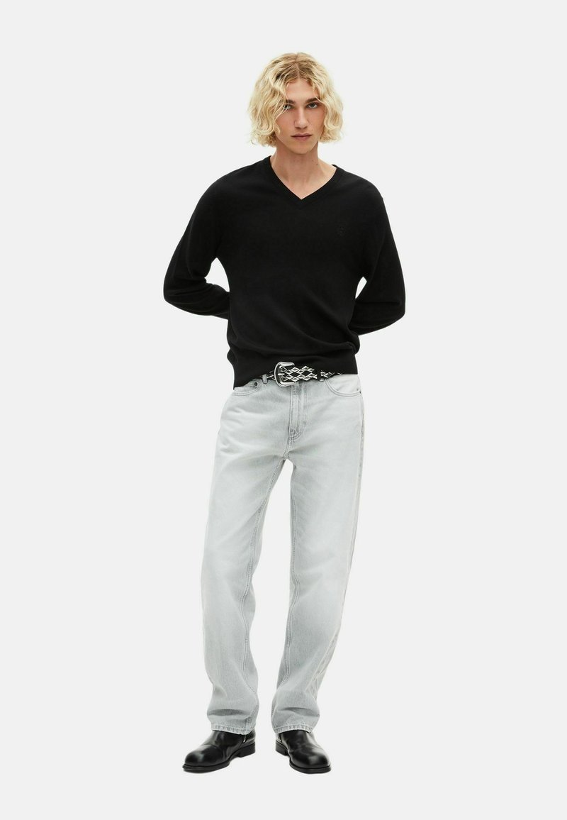 Pull noir à col en V, jean droit gris clair, ceinture à motif et bottes noires. Le mannequin se tient avec les mains derrière le dos devant un fond blanc.