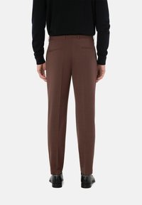 Bruine op maat gemaakte broek van een gestructureerde stof; twee achterzakken en een slim fit ontwerp. Zwarte schoenen worden gedragen, met zichtbare schoenhardware.