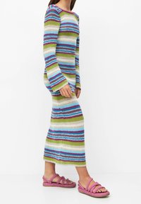 Robe en maille rayée à manches longues dans des tons verts, bleus et violets, assortie à des sandales à brides roses. La texture semble légère et aérée.