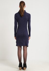 Långärmad, figurnära klänning i djup lila, tillverkad av ett mjukt stickat material. Har en cowl-halsringning och faller precis ovanför knäet. Texturerad tyg.