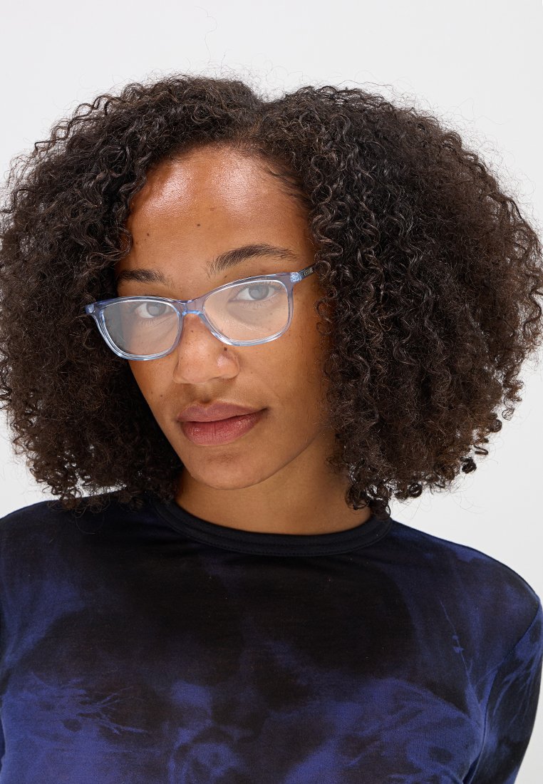 Gafas de sol rectangulares transparentes de color azul con bordes redondeados sutiles. Llevadas por una persona con cabello rizado y oscuro, sobre un fondo blanco liso.
