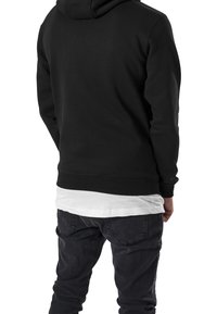 Felpa nera con cappuccio dalla texture liscia, polsini e orlo a costine. Indossata sopra una camicia bianca, abbinata a jeans scuri e consumati.