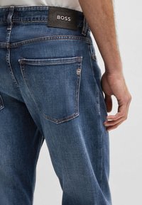 Mörkblå denimjeans har en läderetikett från BOSS i midjan, med synliga sömmar och två bakfickor, varav en med en logodetalj.
