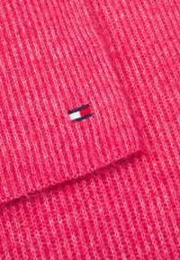 Rosa stickad scarf med ribbad textur. Har en liten broderad logotyp i röd, vit och marinblå. Mjuk material med ett något ludet utseende.