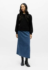 Pull noir à manches longues avec épaules bouffantes, associé à une jupe maxi en denim bleu. Le modèle porte des bottines noires à bout carré et à talons.