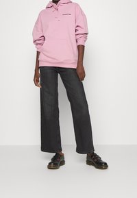 Pink hooded sweatshirt with front pocket, paired with wide-leg dark denim jeans and black platform shoes. Simple text design on the hoodie.