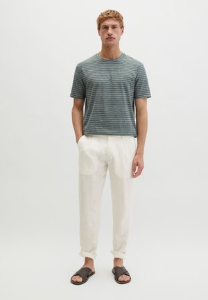 Jonge man staat met handen gedeeltelijk in de zakken, draagt een groen gestreept kort mouw shirt, witte broek en zwarte slippers.