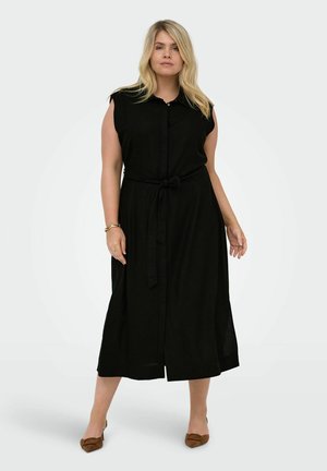 Kvinde står iført en ærmeløs sort midi-kjole med knapper foran og bindebånd i taljen, kombineret med brune spidse sko.