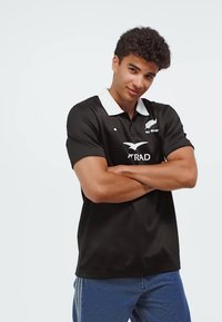 Joven con cabello rizado que lleva una camiseta negra de rugby de los All Blacks y jeans azules, con los brazos cruzados, sonriendo ligeramente, sobre un fondo liso.