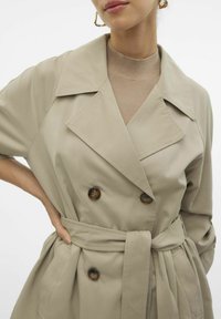 Gabardina beige de doble botonadura con grandes solapas, acentos de botones y un cinturón atado. Hecha de un tejido suave con un corte holgado.