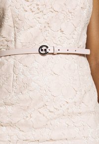 Ljusrosa läderbälte med en cirkulär svart metallspänne, ställt mot ett tyg med spetsmönster och blommiga detaljer.