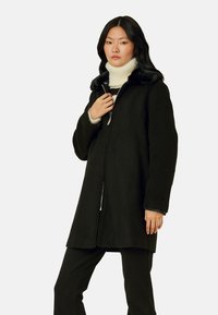 Mustaan villakangastakki faux fur -kauluksella, edessä vetoketjukiinnitys ja löysä malli. Mallilla on päällään vaaleancreme turtleneck. Yksinkertainen muotoilu, ei kuvioita.