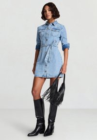 Abito camicia in denim con chiusura con bottoni, maniche corte e cintura annodata in azzurro chiaro. Indossato con stivali neri in pelle al ginocchio e borsa nera con frange.