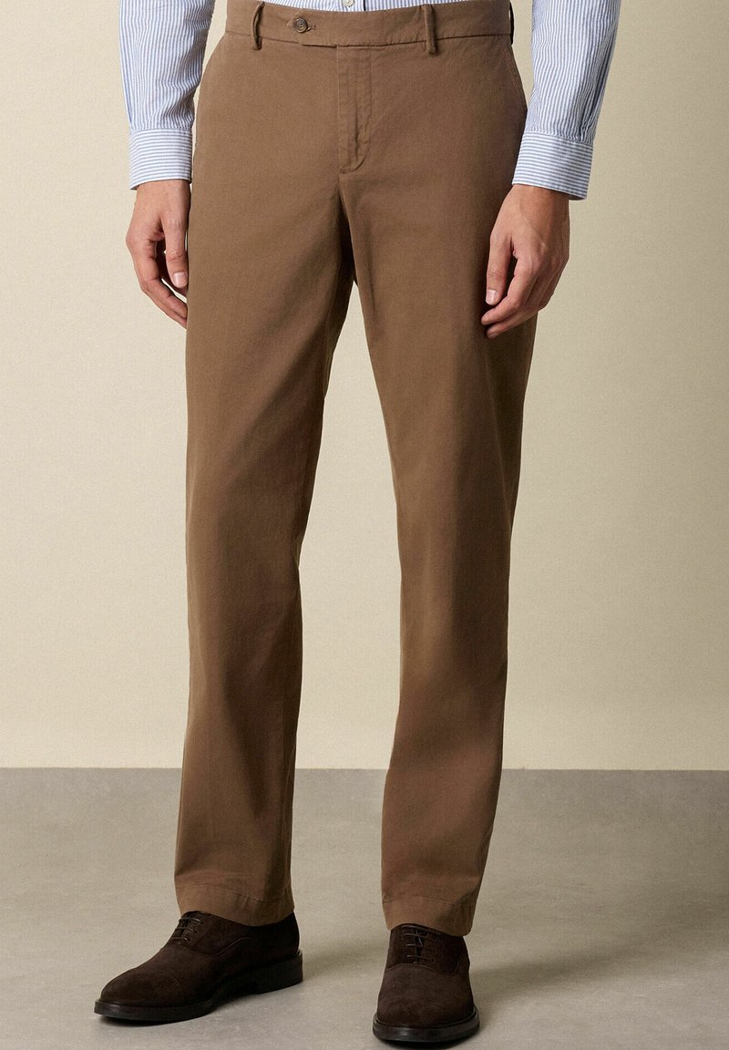 Uomo che indossa pantaloni dritti marroni, scarpe stringate in suede marrone scuro e una camicia blucerchiata a righe azzurre con le maniche giù.