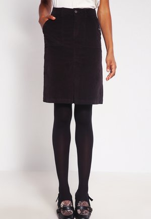 Femme portant une jupe noire longueur genoux avec des poches avant, des collants noirs et des chaussures noires avec dessus à motifs, debout devant un fond blanc.