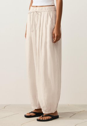 Femeie purtând pantaloni largi, ușor bej deschis, din in, cu talie ajustabilă prin șnur și buzunare, asortați cu papuci negri, stând pe podea placată cu gresie.