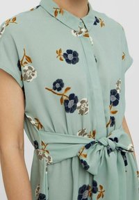 Ljusgrön blus med korta ärmar, blommönster i mörkblått, beige och orange, med krage och knytning i midjan.