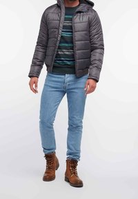 Man med grå vadderad jacka, randig tröja, ljusblå jeans och bruna snörda stövlar som står mot en vit bakgrund.