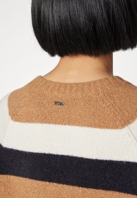 Jersey a rayas con cuello redondo, en colores beige, blanco y azul marino. Textura suave con un detalle de logo en la parte posterior del cuello.
