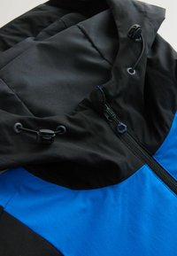 Veste noire et bleue avec fermeture éclair et cordons ajustables à capuche, montrant la texture du tissu et les détails des coutures.