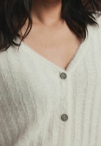 Cardigan blanc à côtes avec un design en V, doté de deux boutons ronds décoratifs ornés d'accents en argent. Tissu doux et texturé.