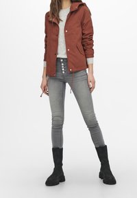 Rust-colored, hooded jacket with button closure and drawstrings; gray sweater underneath; fitted gray jeans; black ankle boots.