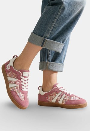Personne portant des baskets à motifs floraux roses et blancs avec des semelles en caoutchouc et un jean bleu retroussé sur un fond uni.