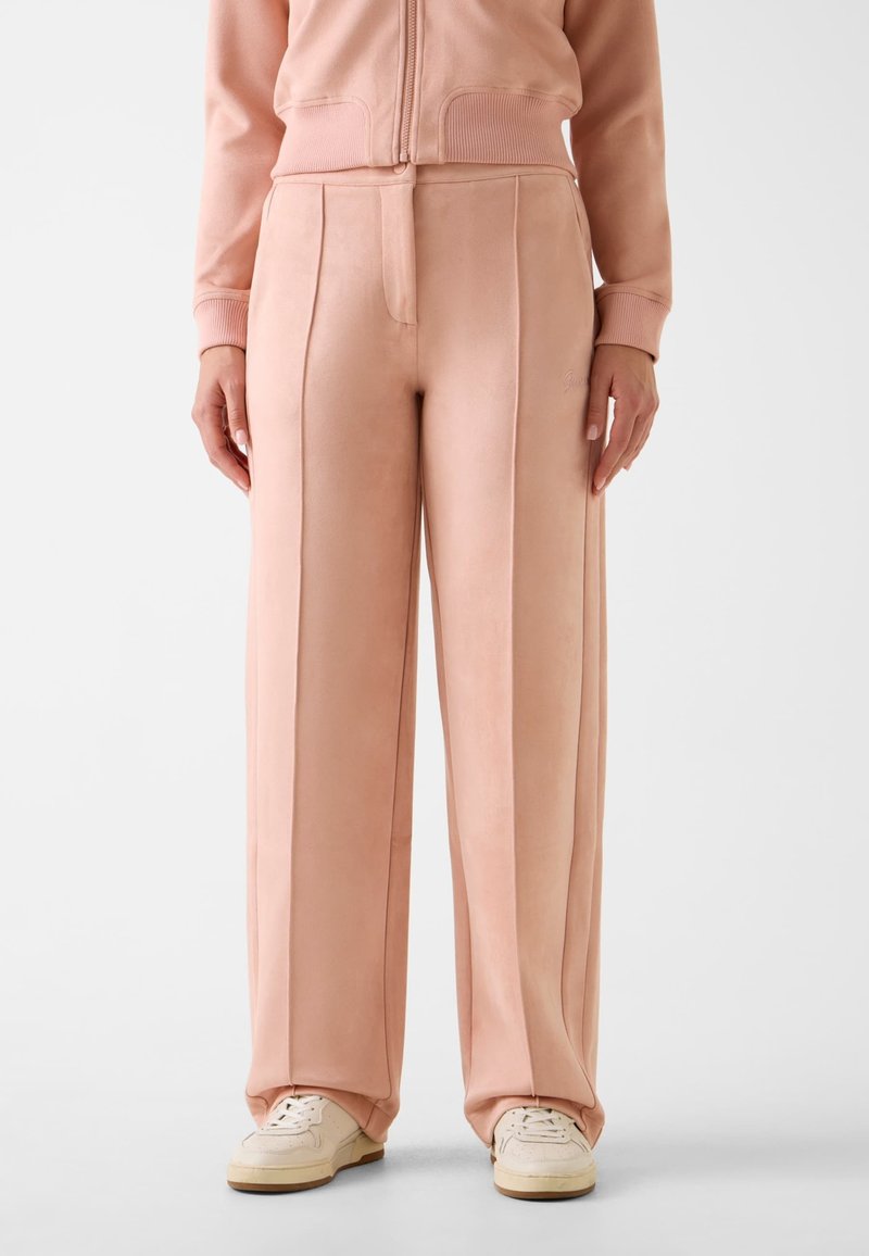 Pantaloni a gamba larga rosa tenue realizzati in tessuto liscio, con pieghe frontali e un logo del marchio discreto sul lato. Abbinati a scarpe da ginnastica bianche.