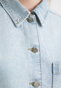 Light blue denim shirt featuring a classic collar, visible stitching, and four metallic buttons with engraved designs. A chest pocket is included.