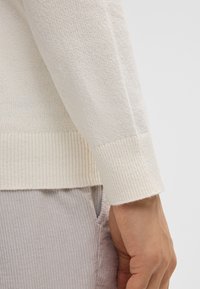 Cuff di maglione lavorato a maglia beige chiaro con texture a coste. Tessuto morbido e design aderente. Pantaloni grigi a coste parzialmente visibili sullo sfondo.