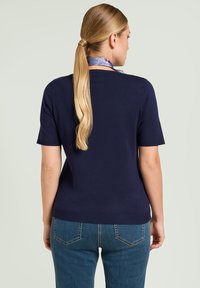 Suéter azul marino de manga corta con escote redondo, cintura ajustada, textura lisa y puños acanalados. Combinado con pantalones vaqueros azules, visto desde atrás.