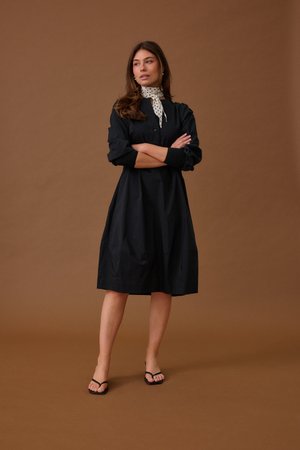 Žena stojící se založenýma rukama v černých šatech po kolena, s bílým puntíkovaným šátkem na krku a černými otevřenými lodičkami na hnědém pozadí.