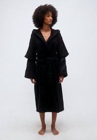 Γυναίκα που στέκεται ξυπόλυτη, φοράει μαύρη ρόμπα με κουκούλα, φαρδιά μανίκια και δεμένη ζώνη στη μέση, σε απλό φόντο.