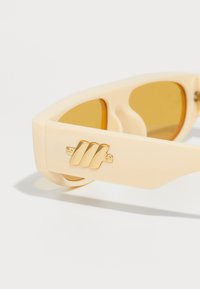 Occhiali da sole color crema con dettagli in metallo dorato e lenti giallo-brown. Montatura angolare con un design distintivo sui lati.