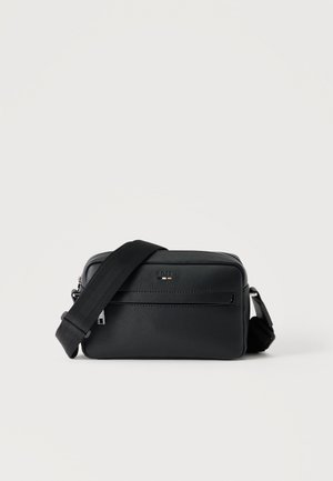 Czarna teksturowana skórzana torba crossbody z przednią kieszenią na suwak i regulowanym szerokim materiałowym paskiem na ramieniu na białym tle.