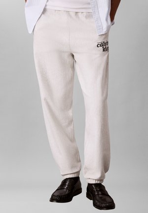 Mees kannab kahvatusvalgeid spordipükse Calvin Klein logo ja mustade nahast loafersitega, käed taskutes, halli ühevärvilise tausta ees.