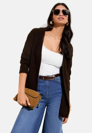 Braune Strickjacke über einem weißen Tanktop, kombiniert mit hochtaillierten blauen Denim-Hosen mit weitem Bein. Hält eine braune Wildleder-Clutch.