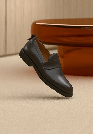 Mocassin en cuir noir à talon bas flottant au-dessus d'un tapis beige, à côté d'une table brillante couleur ambre dans une pièce minimaliste.