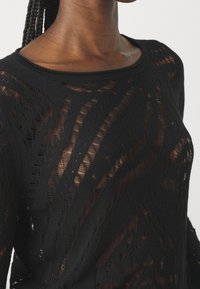 Czarny przezroczysty sweter z misternymi diagonalnymi wzorami koronkowymi, noszony przez osobę z zaplecionymi włosami, odsłaniający szyję i górną część klatki piersiowej.