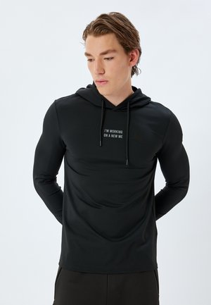 Sudadera negra de manga larga hecha de tela elástica, con un gráfico en el frente que dice "ESTOY TRABAJANDO EN UN NUEVO YO" en blanco, y con cordones ajustables.