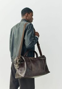 Bolsa de viaje de cuero marrón con dos asas y una correa ajustable. Acabado texturizado con costuras visibles. Cuenta con un bolsillo con cremallera.