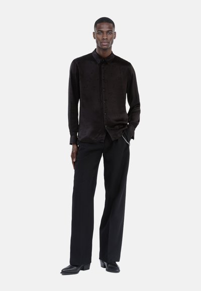 Schwarzes Satin-Hemd mit langen Ärmeln und dezentem Muster, Knopfleiste, kombiniert mit hochgeschnittenen schwarzen Hosen und spitzen schwarzen Stiefeln.