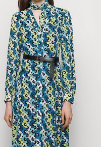 Robe fleurie en bleu, vert et noir, avec un col, des boutons sur le devant et une ceinture noire. Tissu doux avec une texture légère.