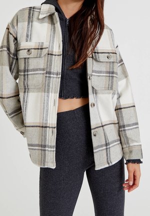 Femme portant une veste boutonnée à carreaux beige et gris sur un pull court zippé foncé et des leggings côtelés sur un fond uni.