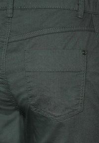 Mörkgrön tygbyxa med en sydd fickdetalj, en mjuk textur och subtil lyster. Inkluderar en knapp vid fickans kant.