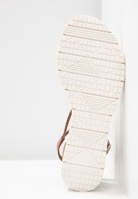 Semelle de sandale plate en caoutchouc blanc texturé, avec des motifs diagonaux et carrés. Brides en cuir marron fixées sur les côtés.